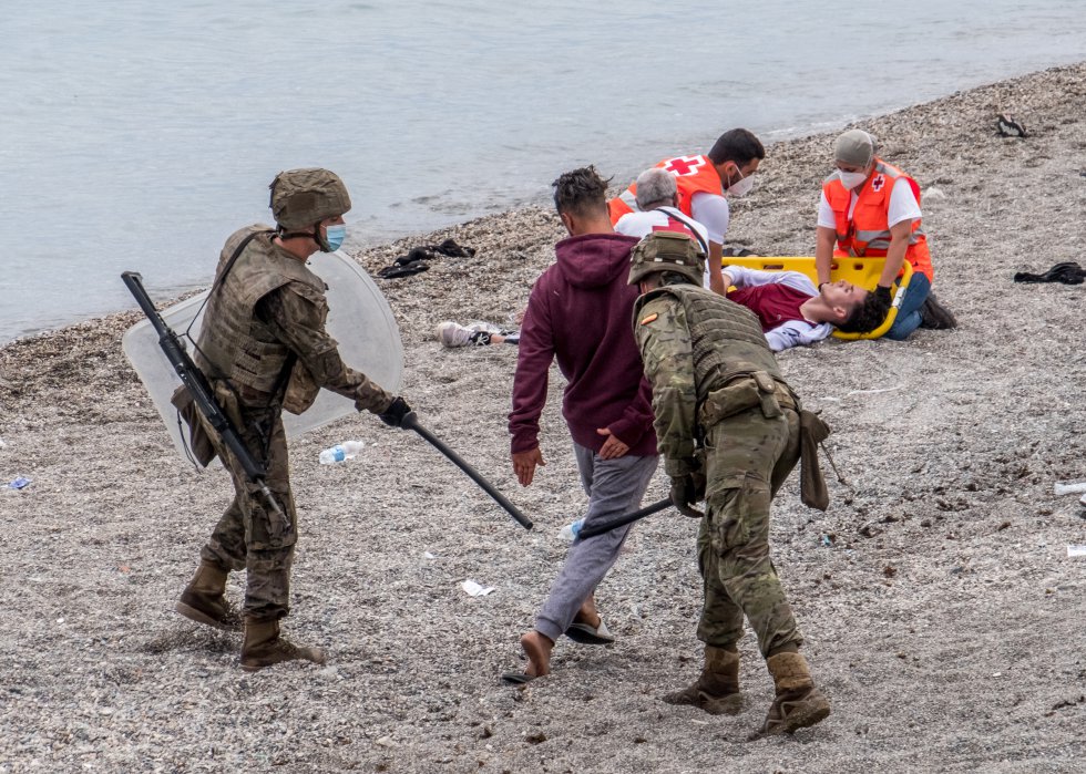 Dos soldados golpean en las piernas a un joven mientras, al fondo de la imagen, dos trabajadores de la Cruz Roja atienden a otro. Varias unidades del Ej&eacute;rcito de Tierra se han desplegado en la madrugada del martes en Ceuta para ayudar en las labores de control de las calles de la ciudad tras la entrada de m&aacute;s de 8.000 inmigrantes, la mayor&iacute;a marroqu&iacute;es, por los espigones fronterizos.